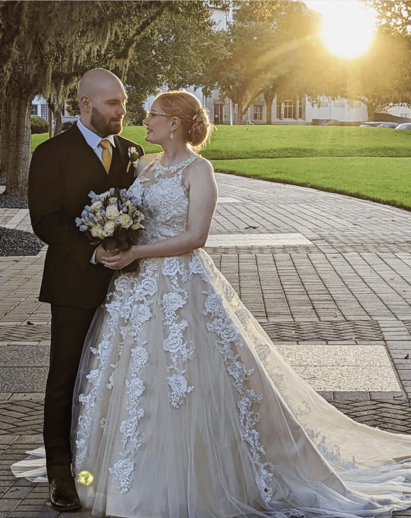 Bride in elegant lace wedding gown holding flowers, glowing in soft sunlight.