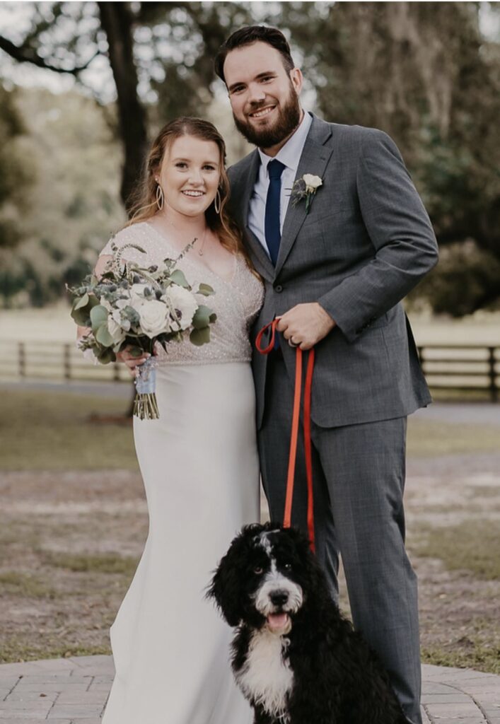 Newlyweds smiling with their black and white dog outdoors.