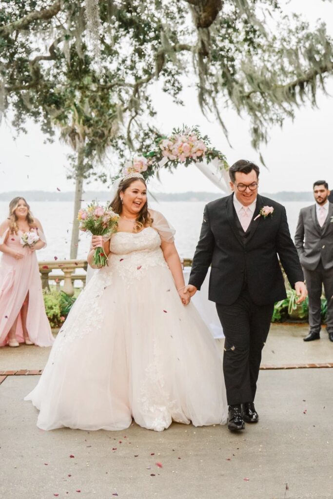 Newlyweds joyfully walk hand in hand outdoors under a floral arch.