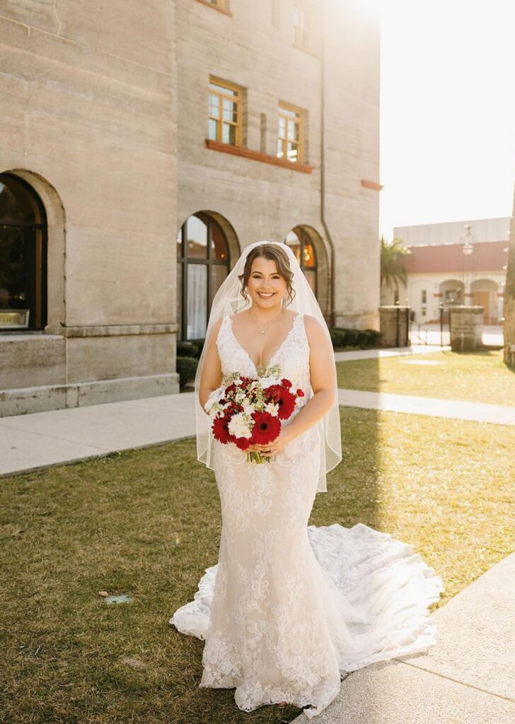 Bride smiling in lace wedding dress holding a red and white flower bouquet.
