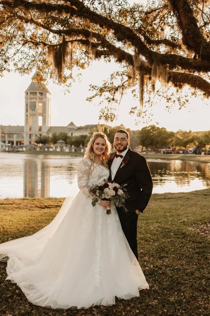 Custom Rachel Ann Bridal gown: bride in an off-the-shoulder ball gown with her groom under oak trees by the water