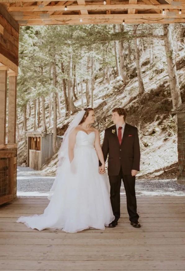 Custom Rachel Ann Bridal gown: bride in a strapless ball gown with her groom in a burgundy suit on a rustic porch