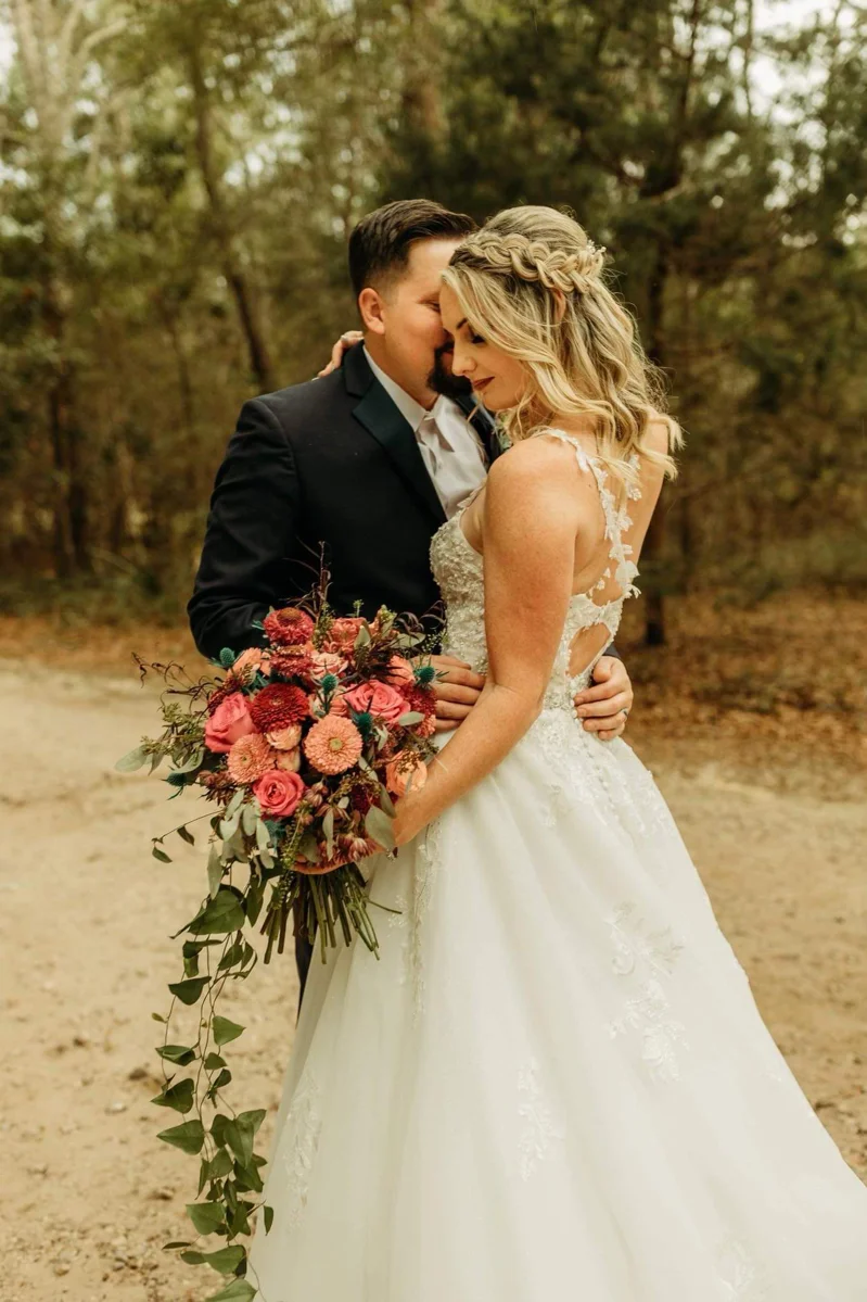 Custom Rachel Ann Bridal gown: bride in a strapless A-line dress with a red and pink bouquet beside her groom in the woods