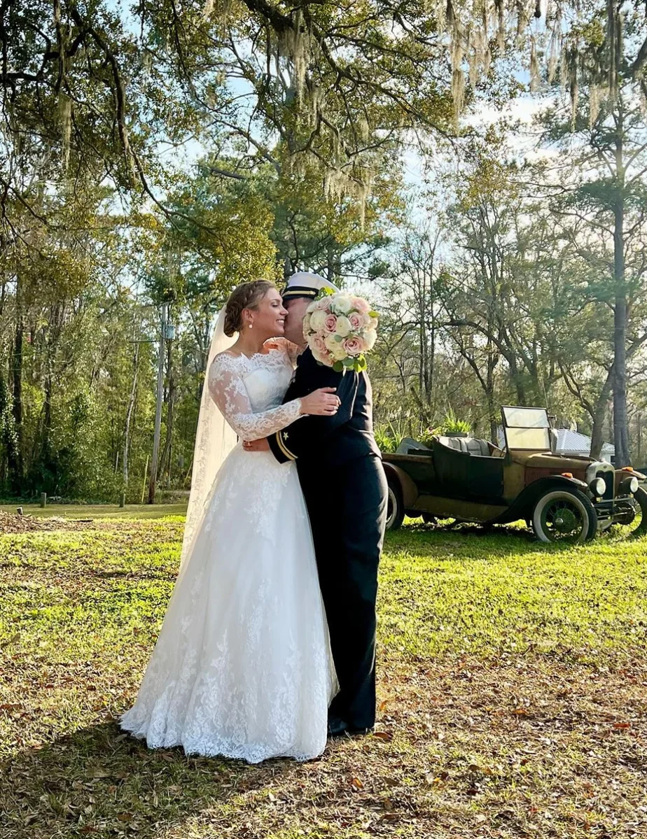 Custom Rachel Ann Bridal gown: bride in a strapless lace dress and veil embracing her groom beside a vintage car