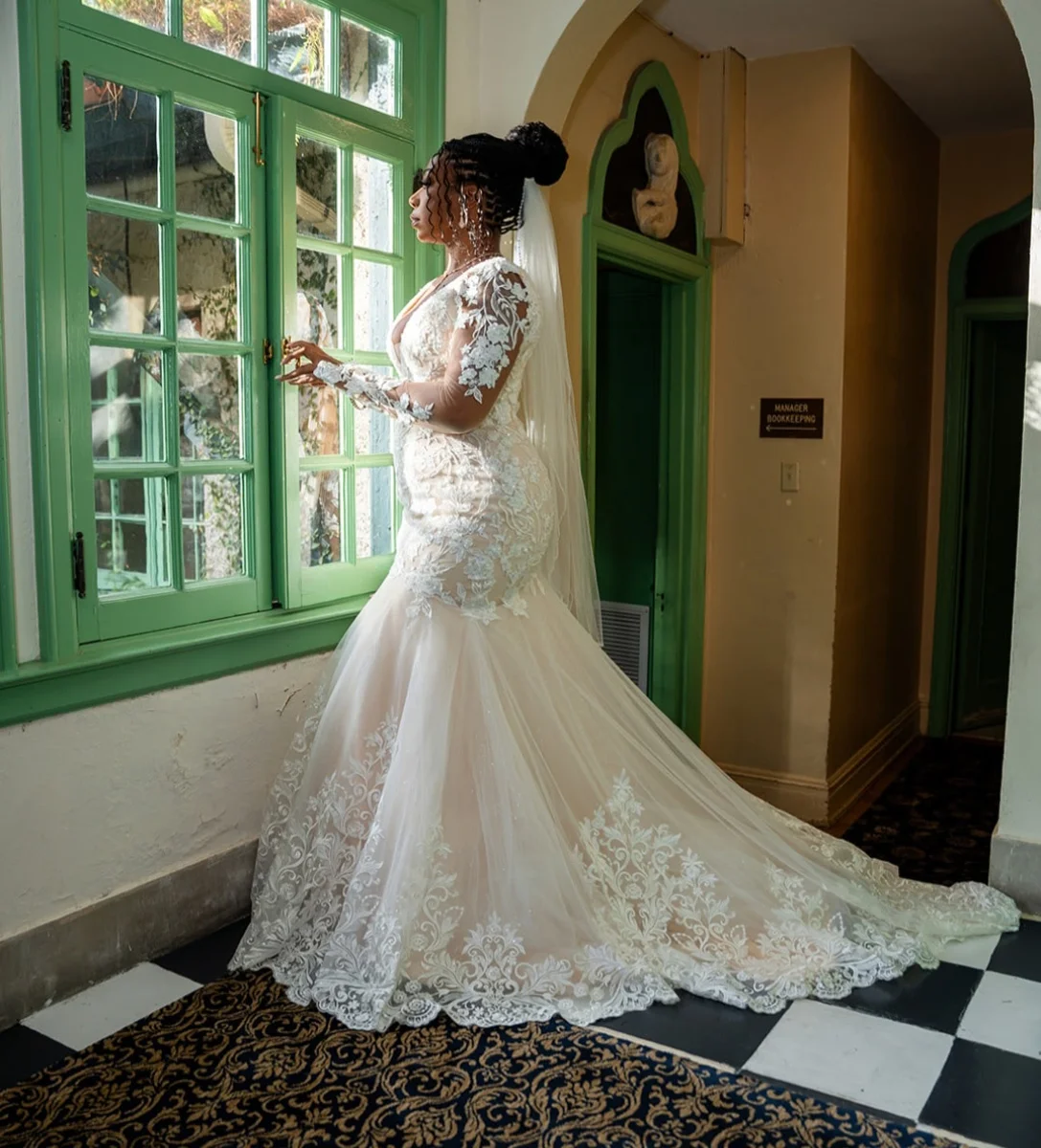 Custom Rachel Ann Bridal gown: bride in a long-sleeve lace mermaid dress beside a green-framed window