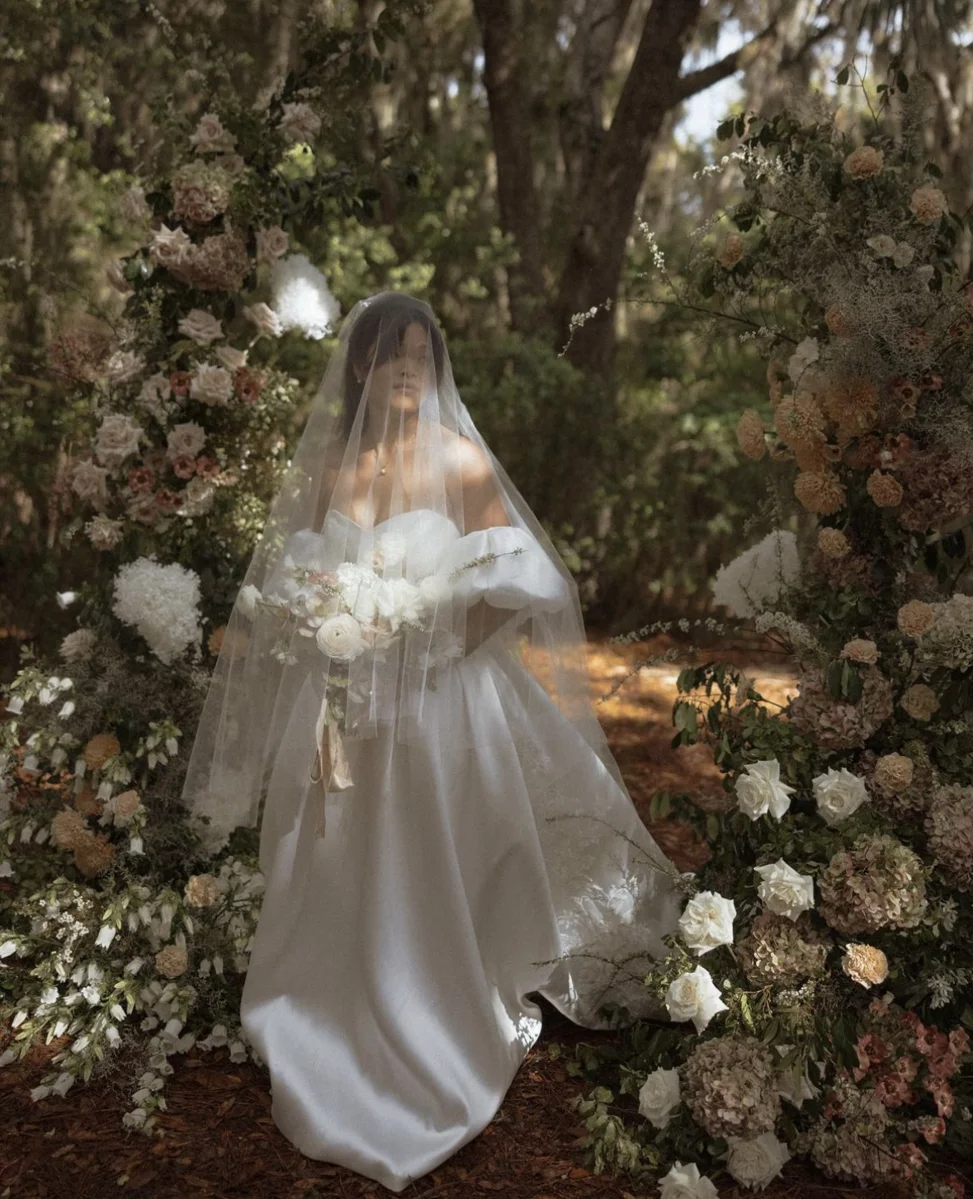 Diane on her wedding day in her custom off-the-shoulder gown and long veil among flowers